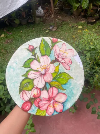A circular hand-painted edible fondant sugar plaque displaying detailed pink apple blossoms, rich green leaves, and delicate buds over a soft watercolor-style blue background, resting on a miniature wooden display easel.