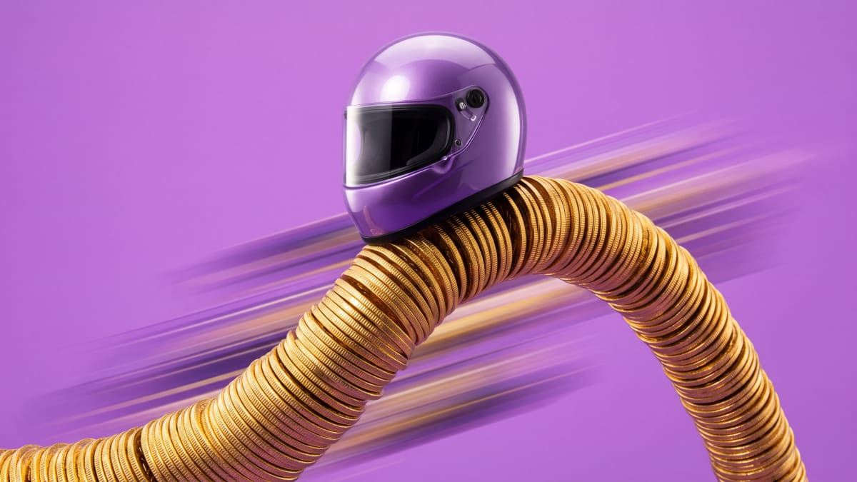 Still life of a racing helmet riding a curving stack of gold coins on a lilac backdrop