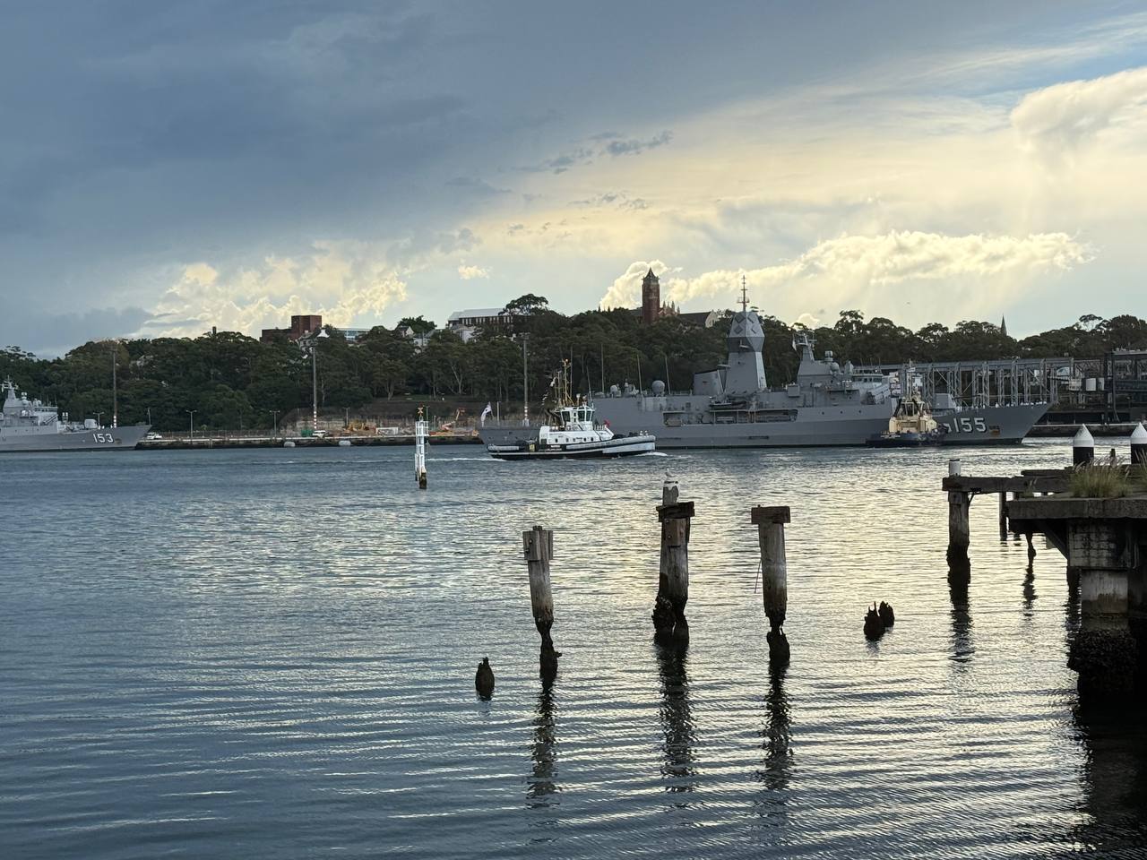 HMAS Ballarat and HMAS Stuart at anchor with tug boat between them, old wooden pier posts in foreground, moody grey sky over Sydney Harbour