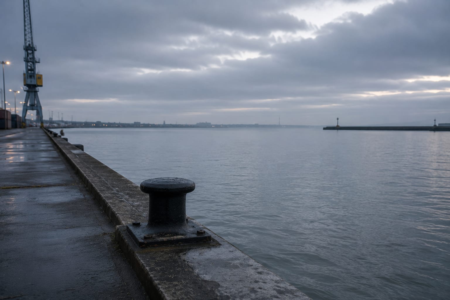 A single bollard on an empty quayside at a UK port with a heavy mooring rope coiled around it, an empty berth stretching beyond, and a single vessel approaching on the grey horizon