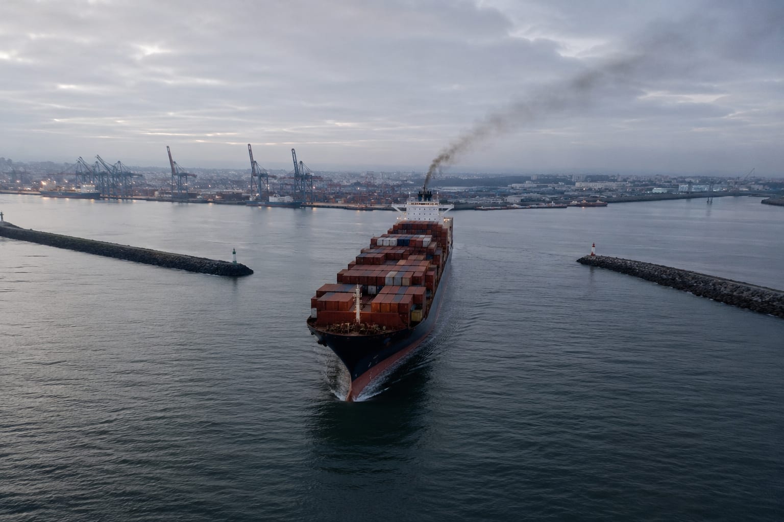 Aerial photograph of a large container ship entering a European port at dawn, passing between breakwaters with a thin exhaust plume trailing from the funnel against an overcast sky