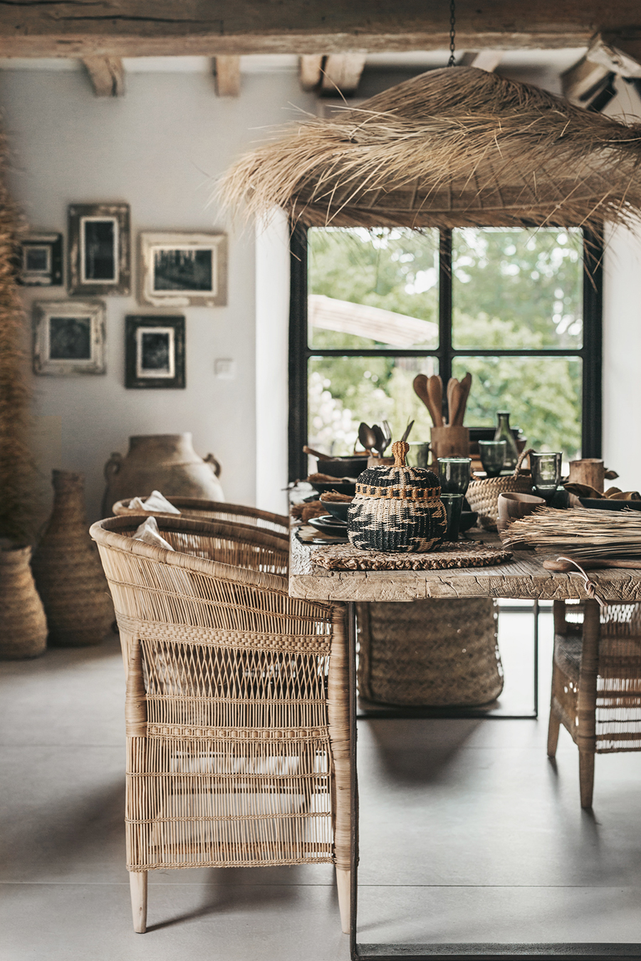 Warm rustic dining space with natural rattan chairs, wooden table decorated with woven baskets and ceramics, thatch ceiling detail, and large window overlooking greenery