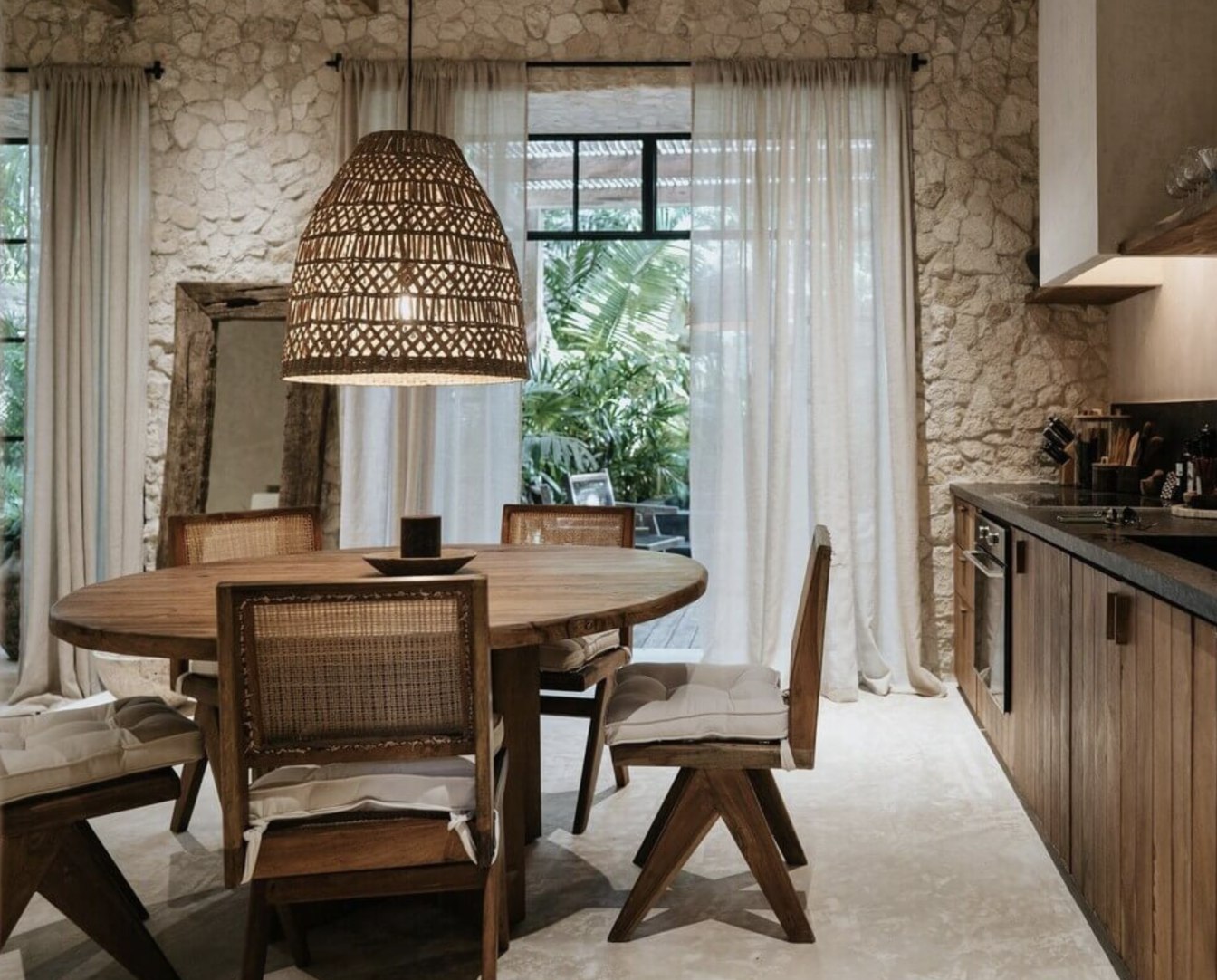 Warm rustic dining space with natural rattan chairs, wooden table decorated with woven baskets and ceramics, thatch ceiling detail, and large window overlooking greenery