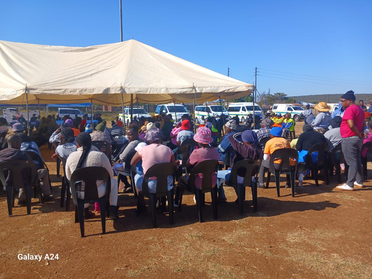 Community members seated under a marquee at an outdoor gathering, with police vehicles parked behind.