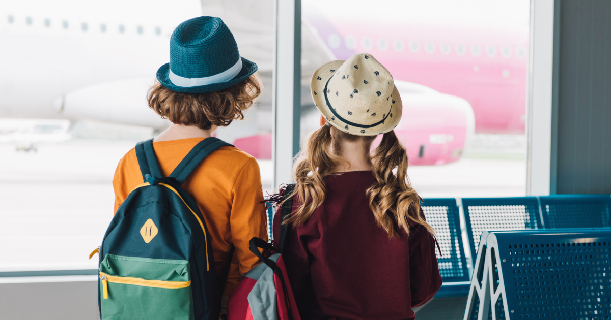 Two children with travel hats and backpacks at an airport terminal window looking out at a pink airplane
