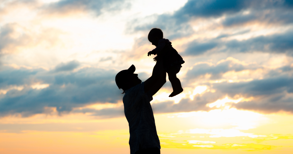 Silhouette of a parent lifting a baby overhead against a dramatic sunset sky