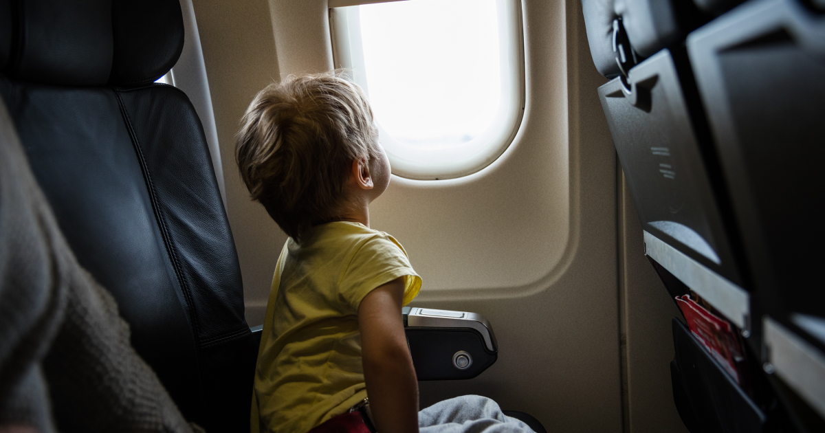Young toddler in a yellow shirt sitting in an airplane seat peering out an oval window