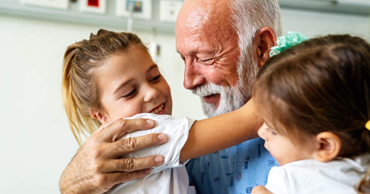Elderly man with a white beard laughing joyfully as two young girls hug him in a hospital setting
