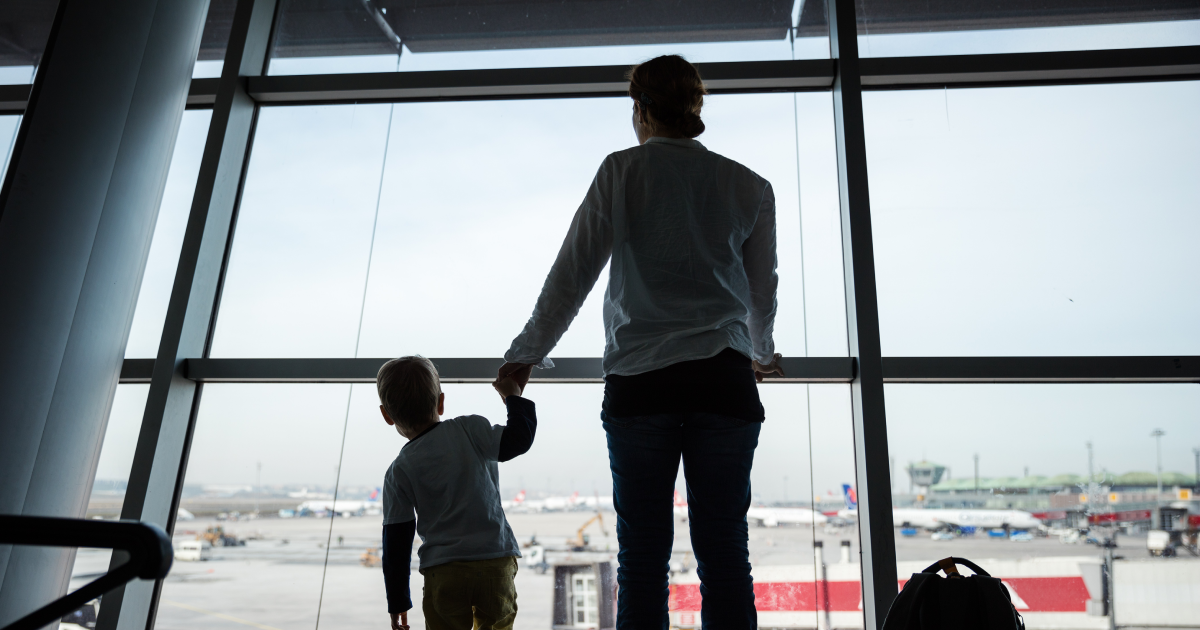 Silhouettes of a parent and young child standing hand in hand at a large airport window overlooking the tarmac