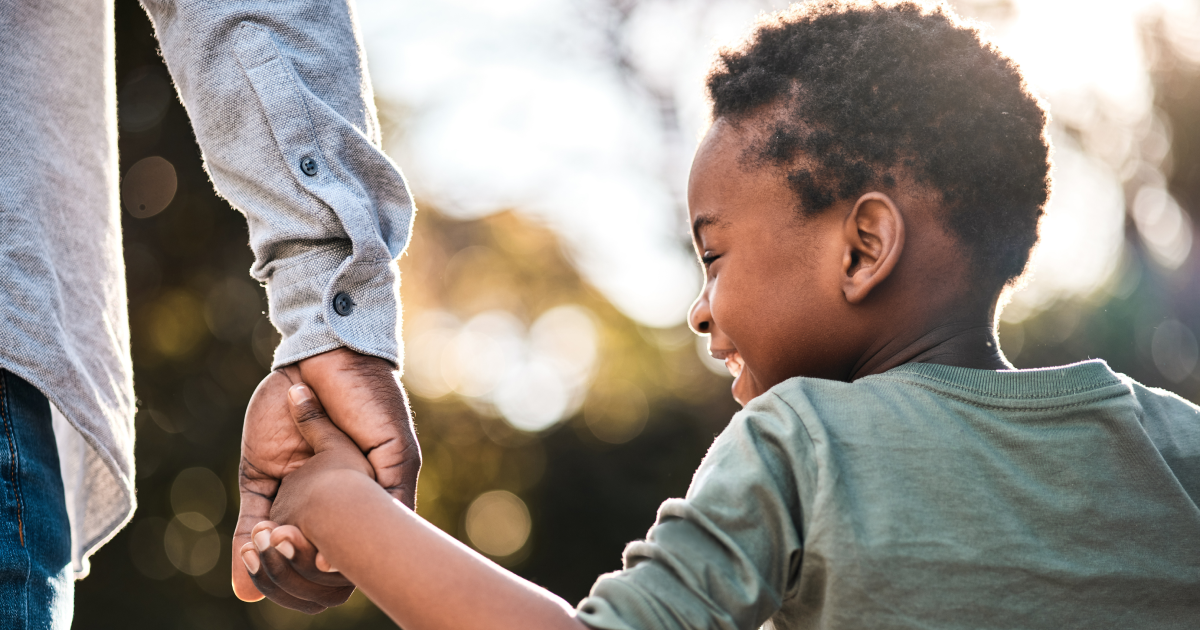 Young boy smiling while holding an adult's hand outdoors with a warm blurred background