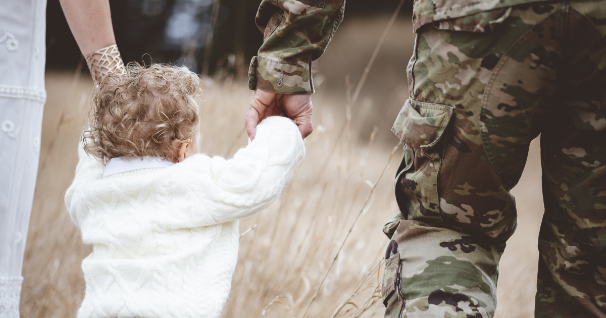 Toddler with curly hair holding hands of an adult in a wedding dress and an adult in military camouflage in a golden field