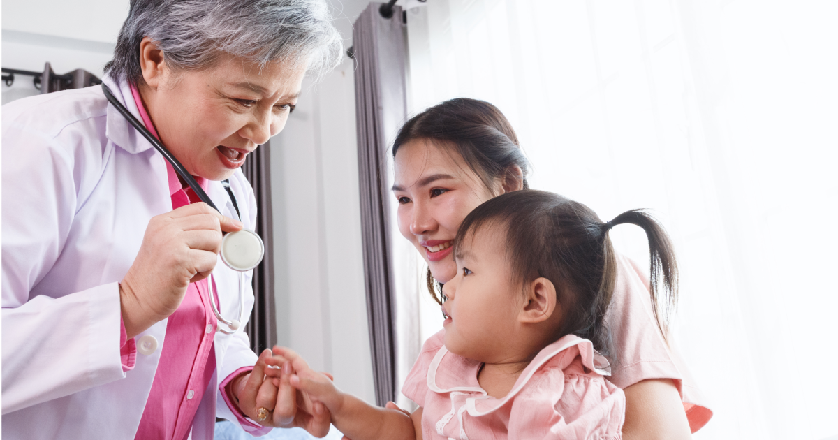 Older female doctor with a stethoscope engaging warmly with a toddler held by a smiling mother during a checkup