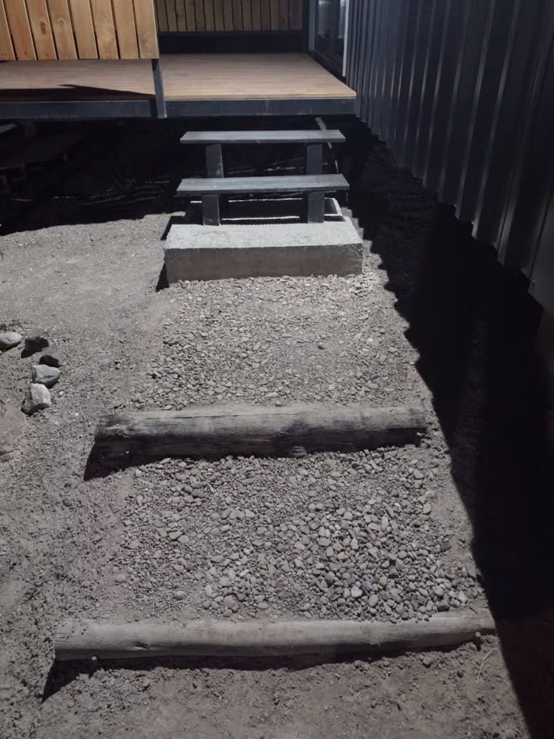 Camino de acceso con escalones de troncos, piedra y luz exterior hacia la cabaña