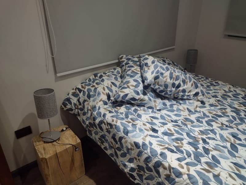 Detalle de sábanas y acolchado de algodón con estampado botánico, almohadas y mesa de luz de tronco