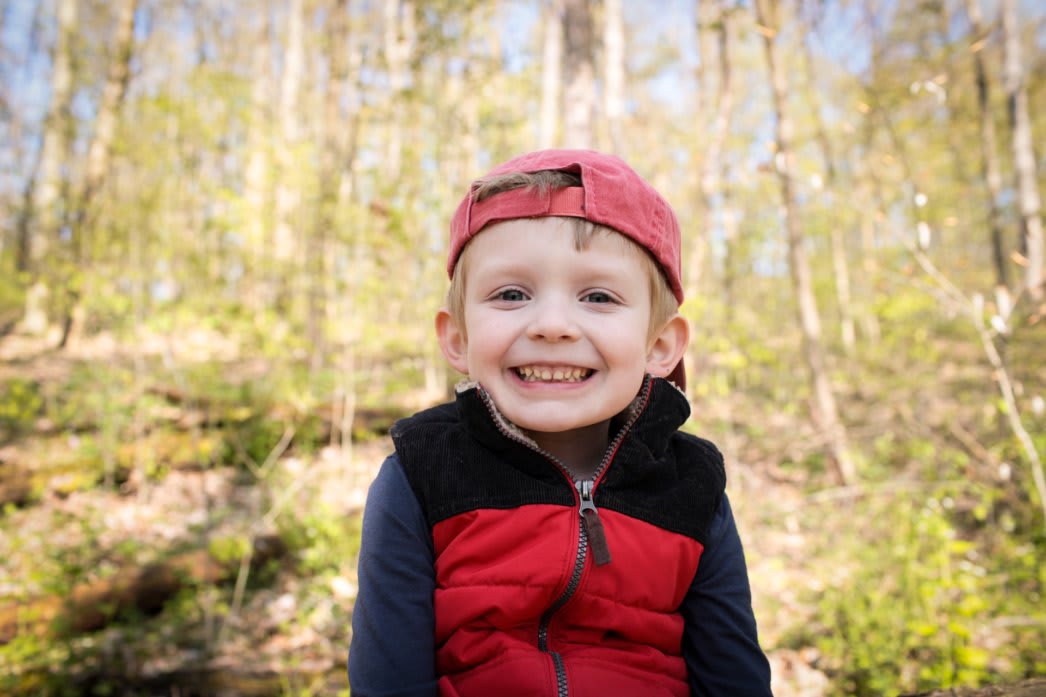 Outdoor photo of Bennett in the woods. Bennett is wearing a red vest, and a red backwards baseball cap, and smiling for the camera.