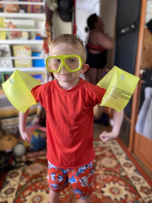 Indoor photo of Bennett in swimming gear. He has floaties, and goggles. He's posing as-if he thinks his floaties are muscles, and he's flexing them. Kate can be seen in the background putting on sunscreen