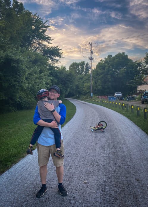 Outdoor photo of a bike trail. In the background a bicycle lays on the ground. Alex is hugging Bennett. Bennett is wearing a bike helmet