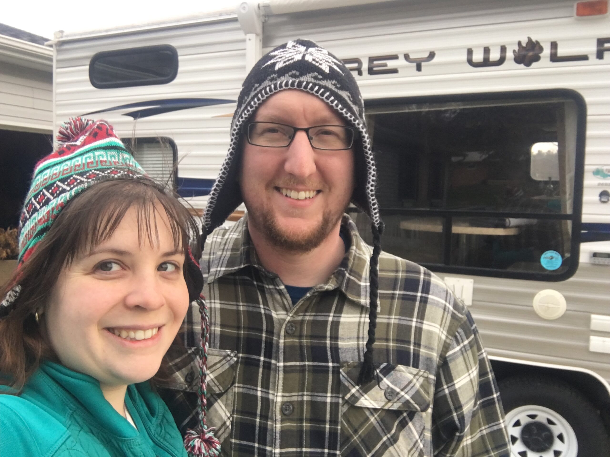 Portrait of Kate and Alex, sitting in-front of their RV. Each are wearing jackets, and hats.