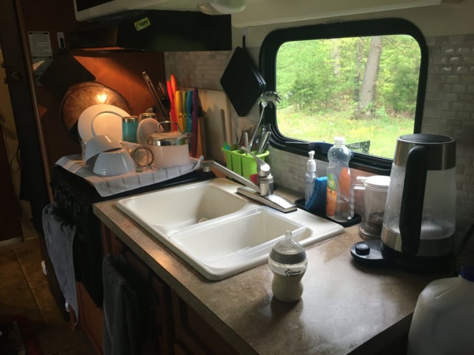 Interior photo of our kitchen area. There is a pile of clean dishes sitting atop the oven, the countertop appears to be recently wiped down, and the sink is empty.