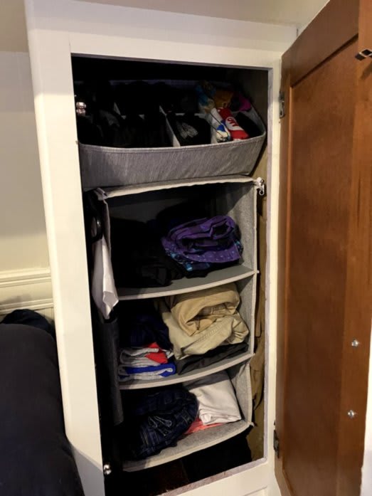 Alex's wardrobe. There is a hanging system that has three shelves. Each shelf has neatly folded articles of clothing. There is a bin on-top of the shelves that holds neatly-folded socks.