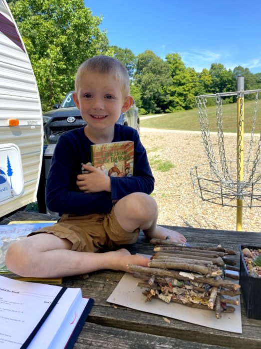 Outdoor photo of Bennett. Ben is sitting on our picnic table, holding a copy of &quot;Little House in the Big Woods&quot;