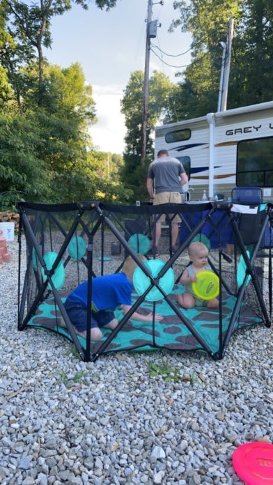 Outdoor photo featuring Devin's playpen. Devin and Bennett are playing together inside. Alex can be seen cooking at his outdoor table in the background.