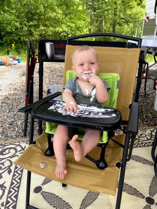 Outdoor Photo of Devin sitting in his folding chair. The chair is attached to full-sized chair. Devin is eating whipped cream, and making a big mess.