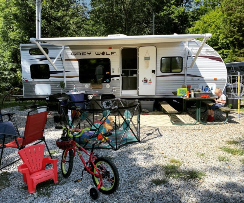 A camper parked on a gravel lot with a playpen, child's bike, and carious chairs set up in front of it.