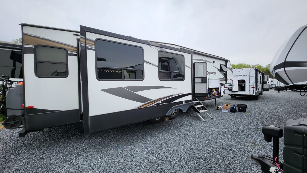 A side view of a large fifth wheel RV. In front of it you can see a much smaller camper with no slides. There is a door open on the rear of the smaller camper and things sitting on the ground as items are being transferred over