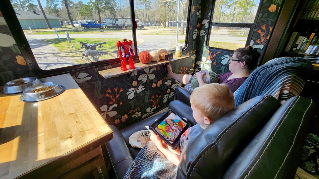 A view out the rear of the camper, which has a large window. Kate, an adult female is winding a ball of yarn while and a blonde haired toddler boy sits in another recliner and looks out the window. They are both sitting in recliners facing the rear window.