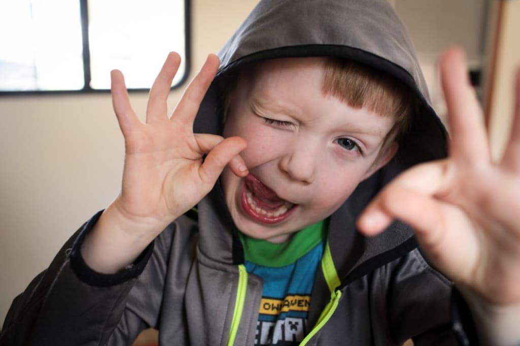 Indoor photo of Bennett. Bennett is wearing a gray jacket with the hood up. He's looking at the camera and making a silly face.