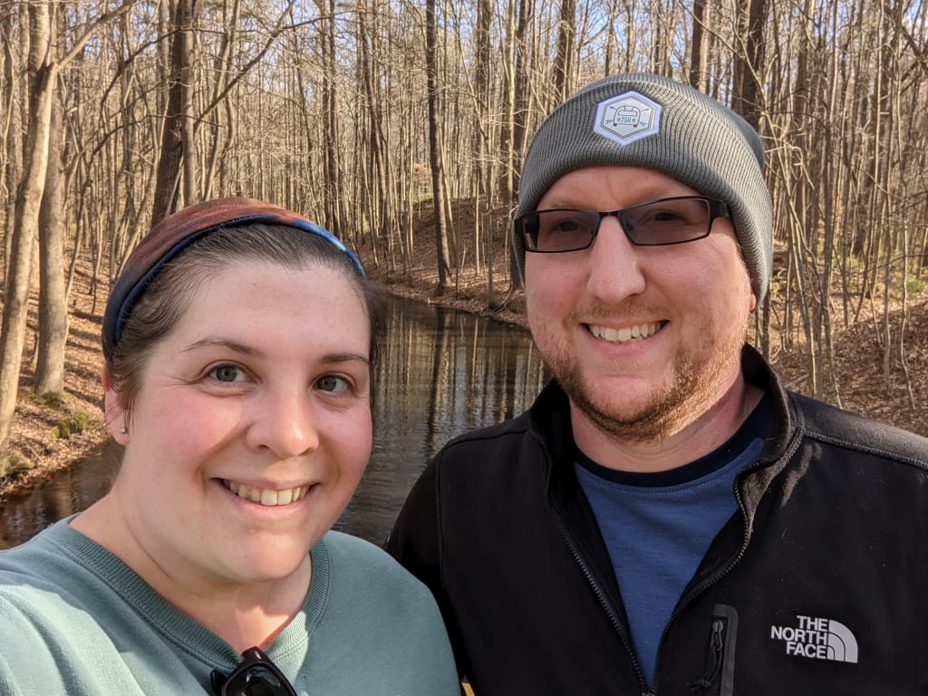 Selfie of Kate and Alex standing beside each other, smiling. In the background there are woods with no green, just brown and dead leaves, and a very black swampy stream.