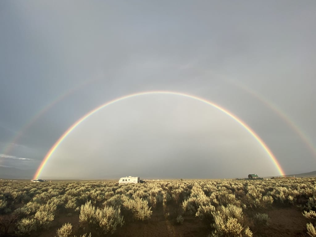 a gigantic vivid double rainbow arching across a blue overcast sky with sage brush in the foreground and a camper parked let of center underneath the rainbow