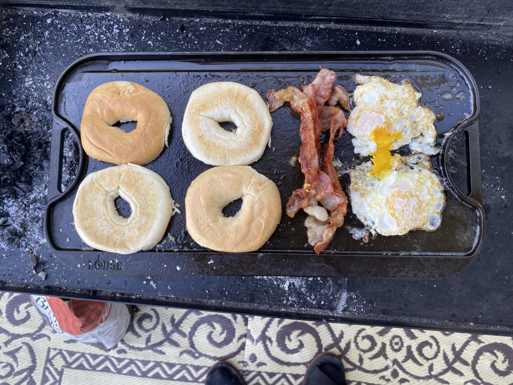 Outdoor photo of Alex's outdoor cooking table. On the table there is a rectangular griddle cooking 2 bagels, bacon, and eggs.