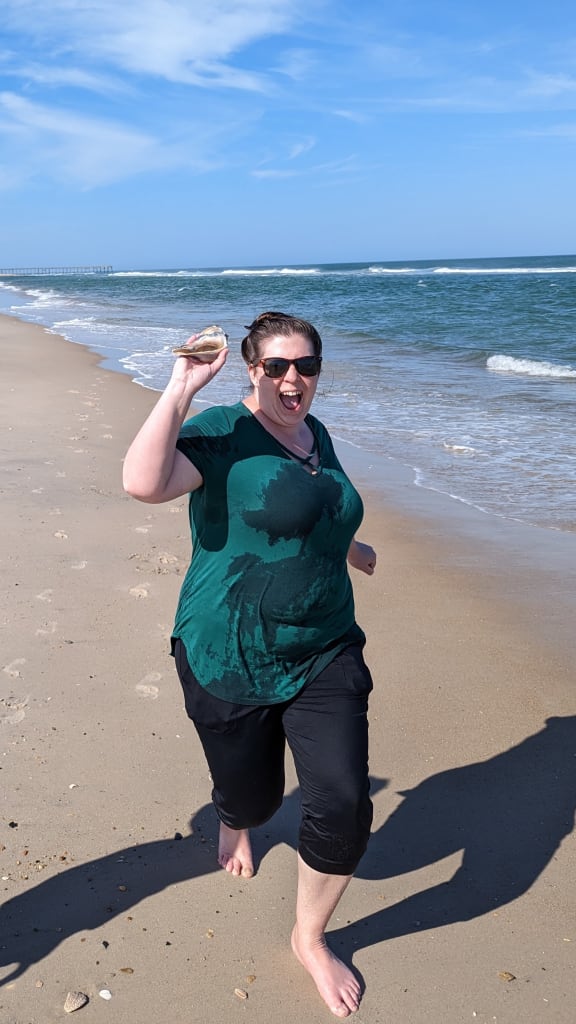 Portrait of Kate at a beach. The waves can be seen crashing in the background.  Kate is wearing a green shirt and pants, and her clothes are soaked. She is making a very excited face and holding a fully intact conch shell.