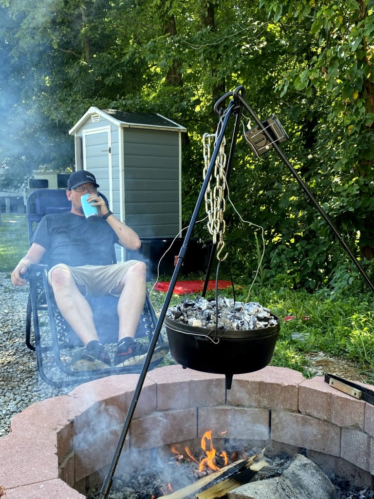 A dutch oven hanging on a tripod over a campfire. Alex can be seen lounging in a chair, drinking from a cup in the background