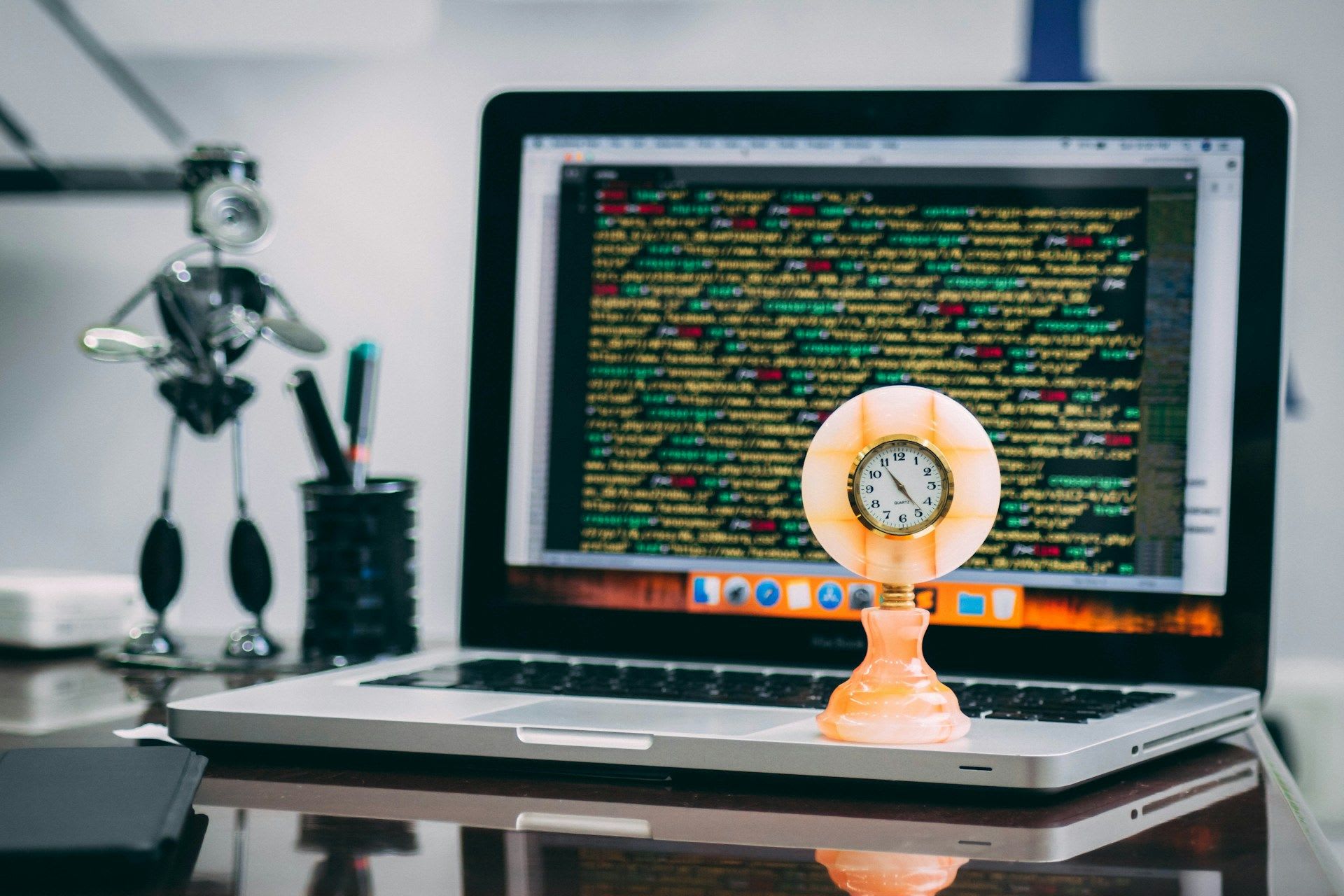 Clock on a desk in the foreground, with a computer screen displaying programming code in the background.