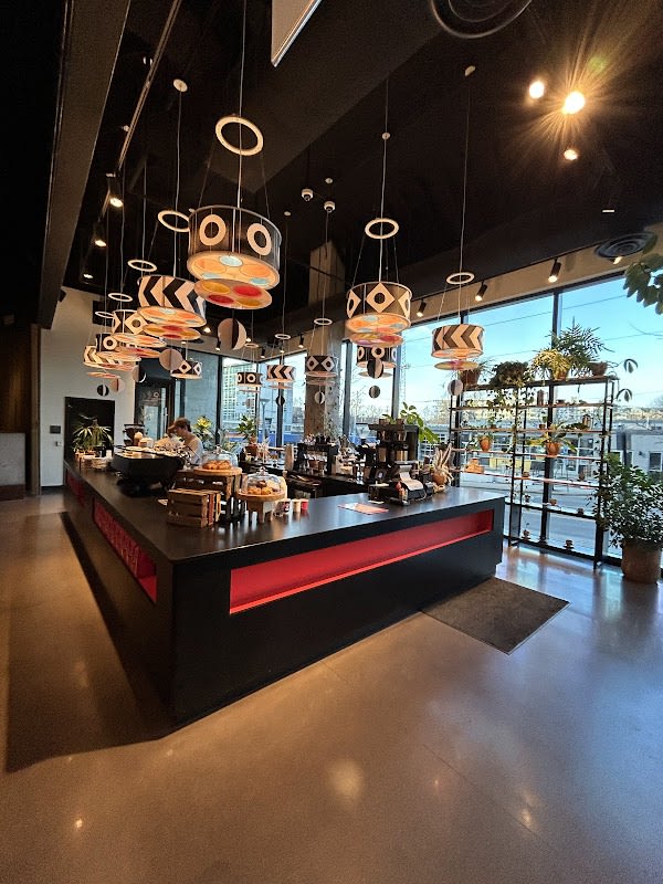 Spacious modern cafe interior with a black service counter lit by red accent lighting, colorful patterned pendant lamps overhead, floor-to-ceiling windows and potted plants