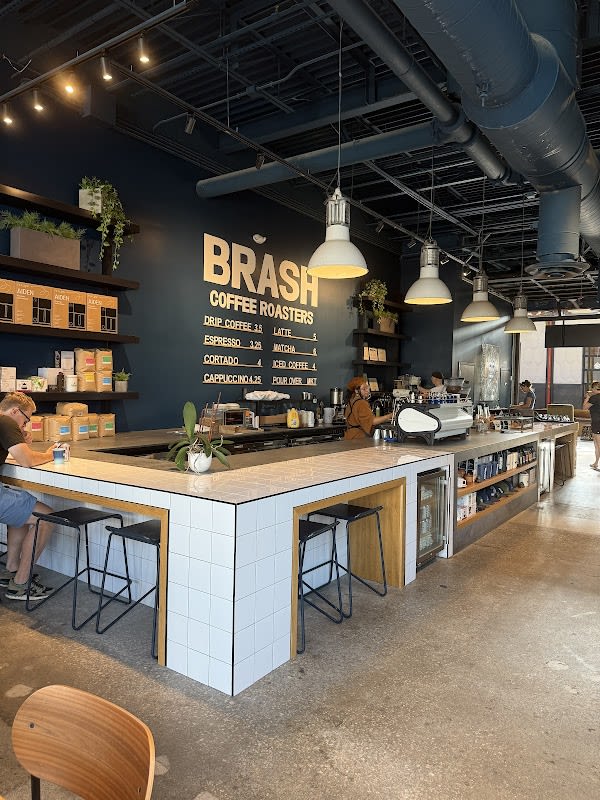 Interior of Brash Coffee Roasters showing a white tiled coffee bar, hanging pendant lights, exposed dark ductwork, and a menu wall listing drip coffee, espresso, cortado, cappuccino, latte, matcha, iced coffee, and pour over