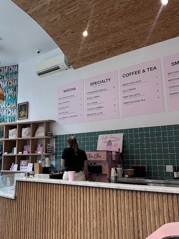 Bright modern matcha cafe interior with pink menu boards listing matcha, specialty and coffee drinks, green tiled counter wall, fluted wood counter and a barista at the register