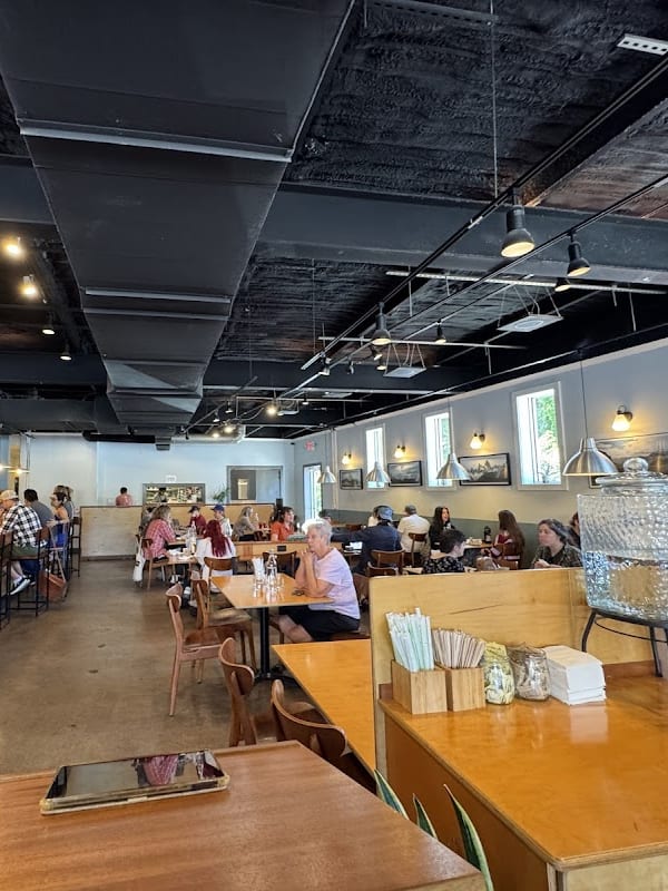 Interior view of a busy cafe with black industrial ceiling, wooden tables, bar seating, and customers enjoying drinks and conversation