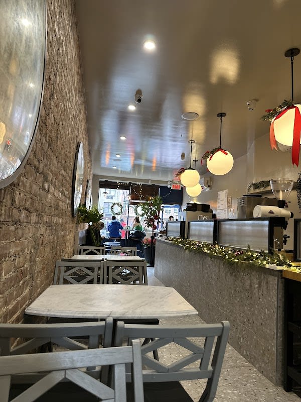 Interior of a cozy cafe featuring exposed brick walls, marble-top tables, gray chairs, and a counter decorated with holiday garland and string lights