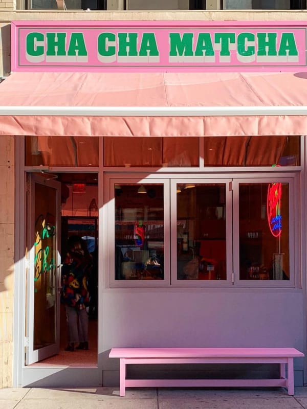 Pink storefront of Cha Cha Matcha cafe with green lettering, pink awning, and pink bench on sidewalk