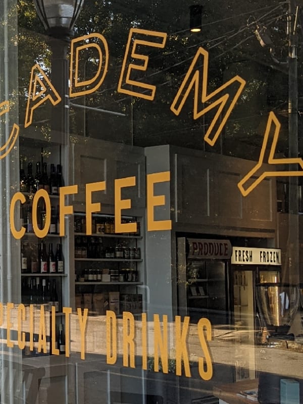 Glass storefront of Academy Coffee with gold painted lettering reading 'Academy Coffee' and 'Specialty Drinks', an Open sign hanging in the door, and shelves of wine and pantry goods visible inside