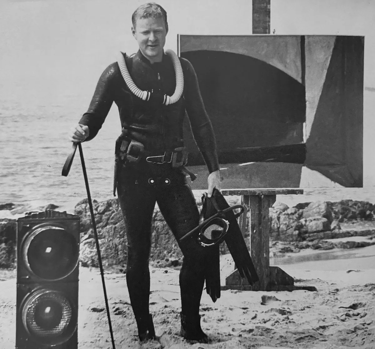 Roger Kuntz in diving gear on the beach next to one of his paintings