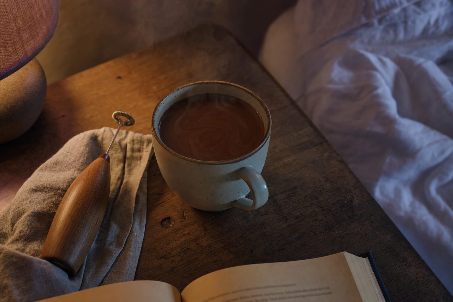 A small ceramic mug of dark caramel-chocolate cocoa on a wooden bedside table at evening, with a wooden frother and book beside it