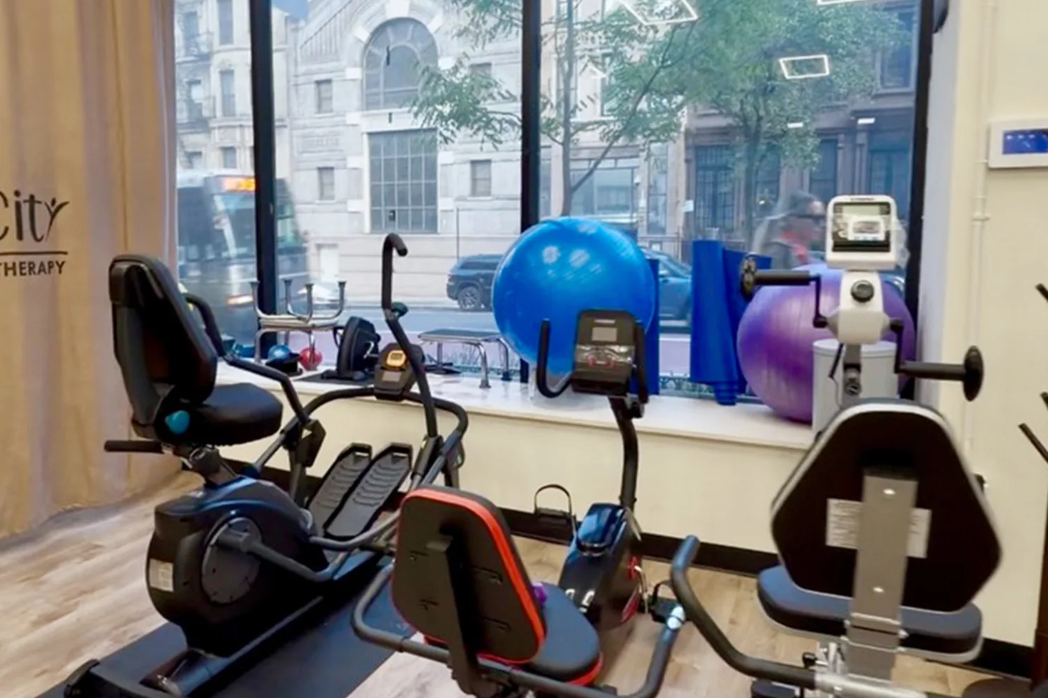Interior of PT of the City physical therapy clinic featuring rehabilitation equipment, stationary bikes, and large windows overlooking the NYC street.
