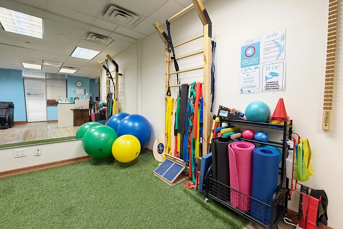 Pediatric therapy gym at PT of the City with colorful equipment including exercise balls, agility ladder, cones and resistance bands on turf flooring