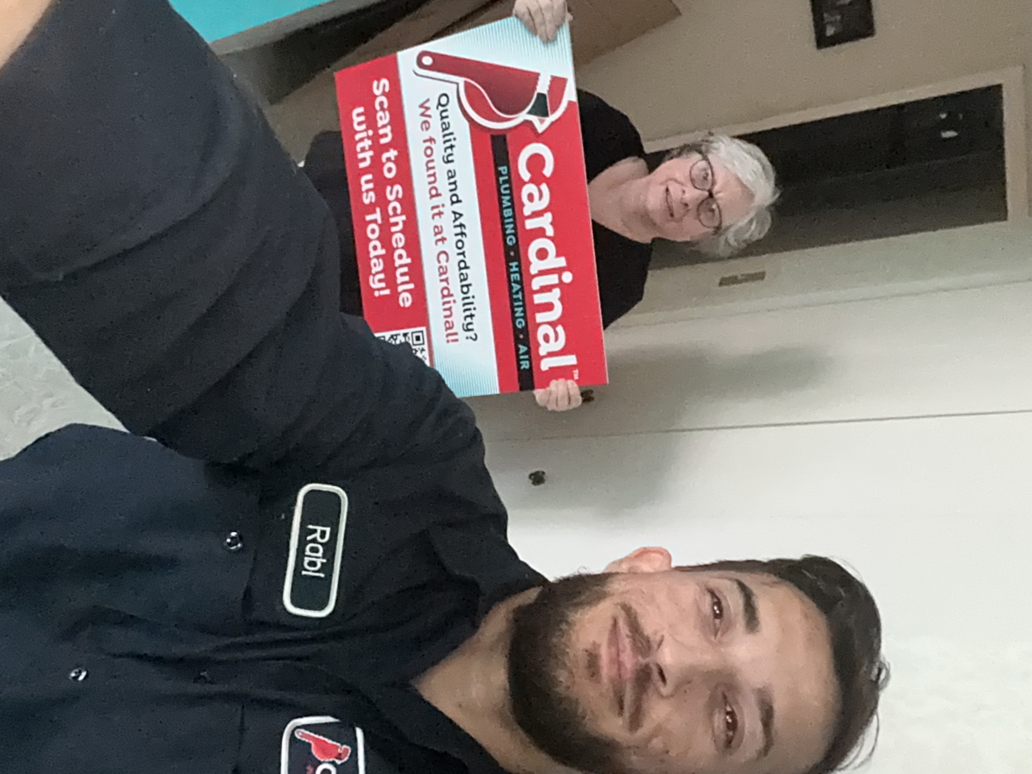 A satisfied homeowner proudly displays our sign while a Cardinal Plumbing Heating & Air Inc technician finishes the job, showing trustworthy service.