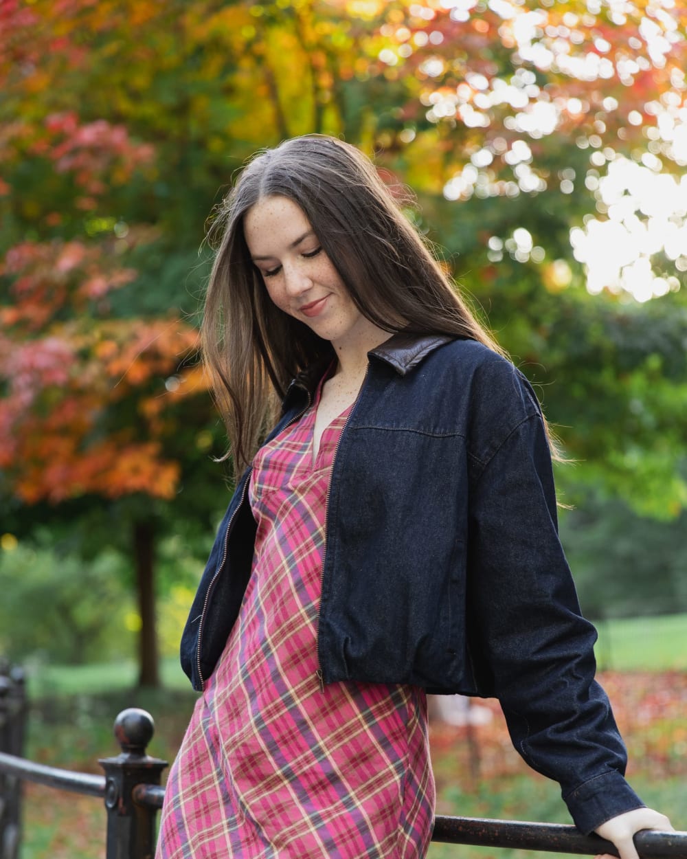 A woman in a plaid dress leaning on a railing in front of autumn trees